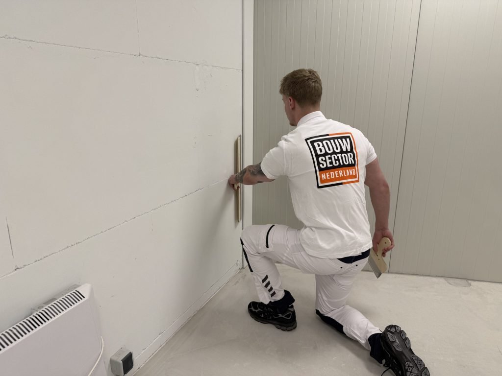 Geknielde stukadoor trekt met een spaan het onderste deel van een gipswand glad, vlak boven de vloer